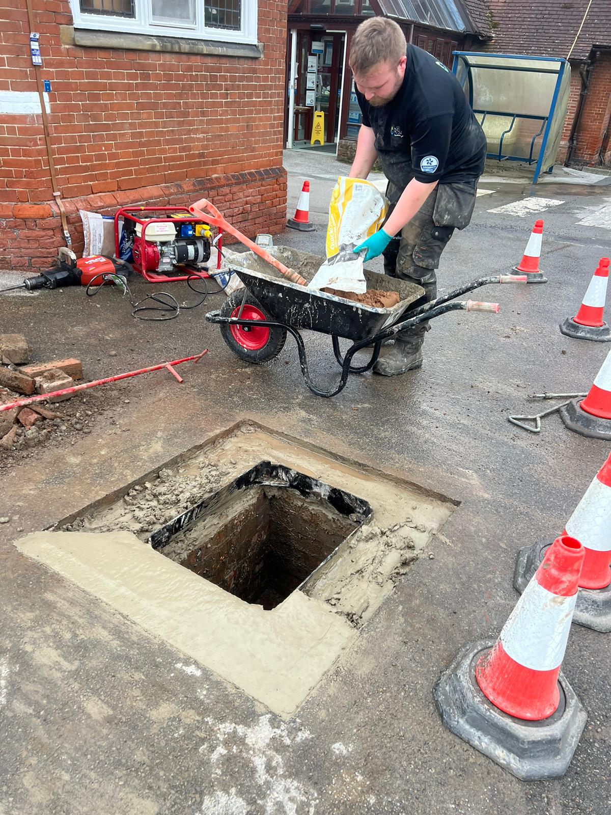 engineer pouring building material into a wheelbarrow in front of an open manhole